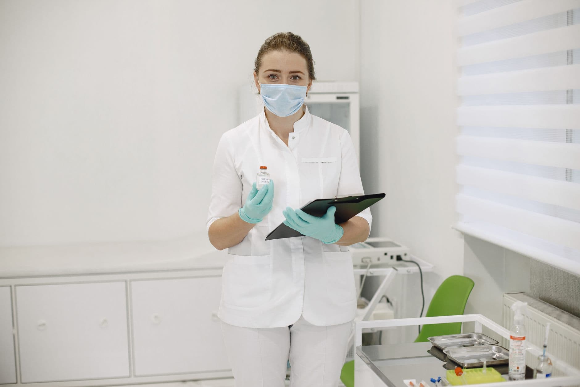 Medical professional in protective gear holding vaccine vial and clipboard in a clinical setting.
